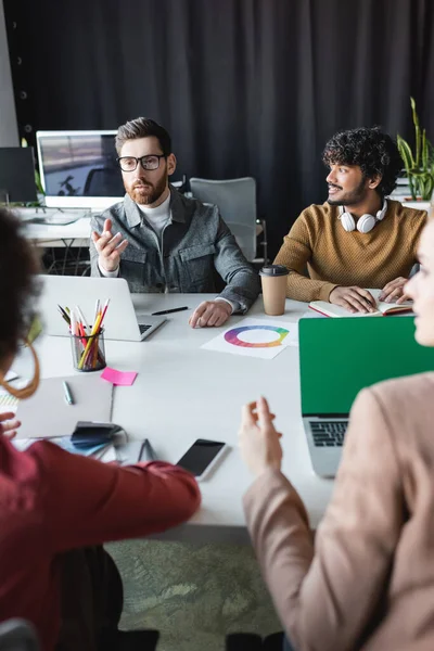 Man in eyeglasses pointing at blurred colleagues near smiling indian man in advertising agency — Stock Photo