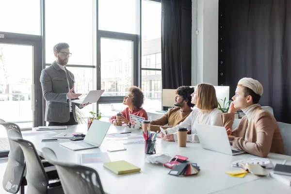 Man with laptop talking to multiethnic advertising managers during meeting — Stock Photo