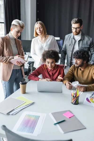 Smiling african american woman pointing at laptop near multiethnic colleagues in advertising agency — Stock Photo