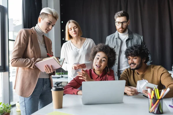 Mujer afroamericana hablando con diseñadores multiétnicos cerca del ordenador portátil en la agencia de publicidad - foto de stock