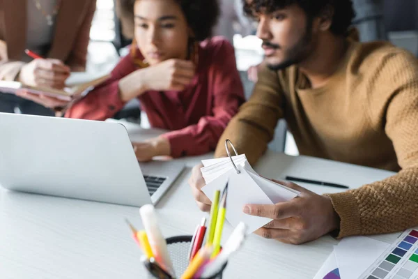 Verschwommene multiethnische Designer arbeiten in der Nähe von Laptop in Werbeagentur — Stockfoto