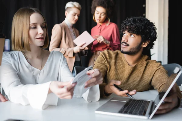 Curly indian man pointing at laptop near colleague and interracial advertising managers on blurred background — Stock Photo