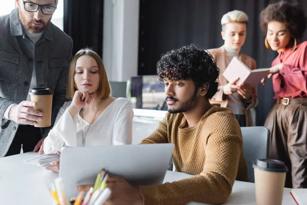 Man with paper cup pointing at laptop near indian colleague in advertising agency — Stock Photo