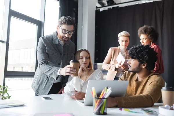 Sonriente hombre con taza de papel apuntando a la computadora portátil cerca de los administradores de publicidad interracial - foto de stock