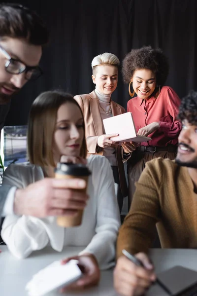 Lächelnde Frauen, die in Notizbüchern neben verschwommenen Kollegen in Werbeagenturen suchen — Stockfoto