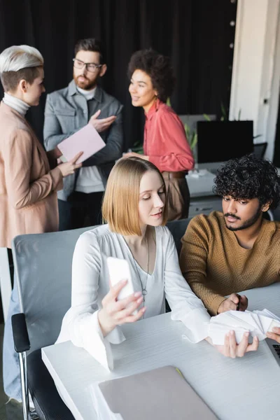 Indio hombre apuntando a las muestras cerca de colega y equipo interracial hablando sobre fondo borroso - foto de stock