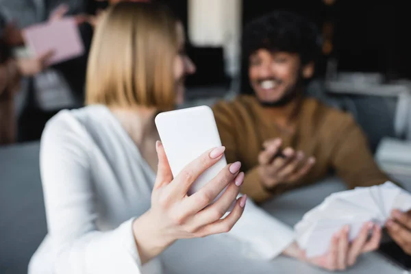 Foco seletivo de smartphone na mão de mulher perto de homem indiano sorrindo no fundo embaçado — Fotografia de Stock