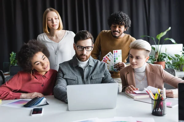 Uomo in occhiali che lavora al computer portatile vicino sorridente colleghi interrazziale in agenzia pubblicitaria — Foto stock