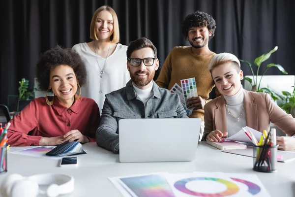 Happy multiethnic advertising designers looking at camera near laptop and color samples — Stock Photo