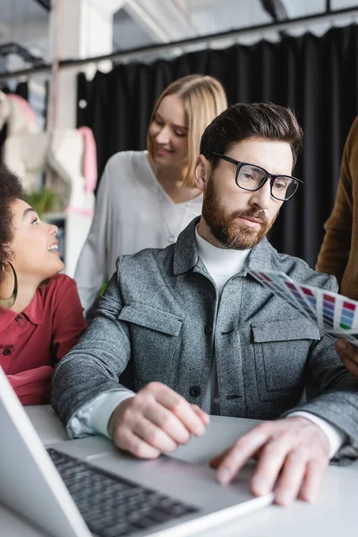Bearded designer working at laptop near smiling interracial women talking on blurred background — Stock Photo