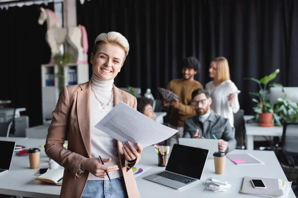 Diseñador alegre con papeles y pluma mirando la cámara cerca de colegas multiétnicos en la agencia de publicidad - foto de stock