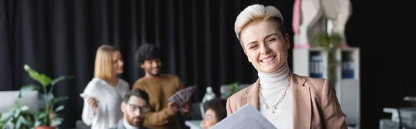 Designer alegre olhando para a câmera perto de colegas multiculturais borrados na agência de publicidade, banner — Fotografia de Stock