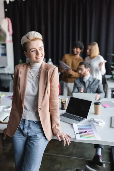 Complacida mujer rubia sonriendo cerca de colegas interracial sobre fondo borroso en agencia de publicidad - foto de stock