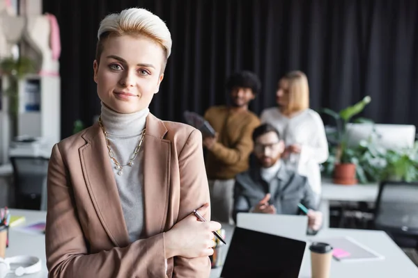 Fröhliche Managerin mit Stift, die in die Kamera schaut, in der Nähe eines verschwommenen interrassischen Teams einer Werbeagentur — Stockfoto