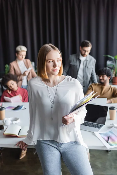 Frau mit Ordnern, die in der Nähe von verschwommenem Laptop und multikulturellen Kollegen wegschauen — Stockfoto