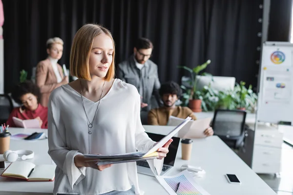 Lächelnde Frau schaut in Ordner neben verschwommenem interrassistischem Team, das in Werbeagentur arbeitet — Stockfoto