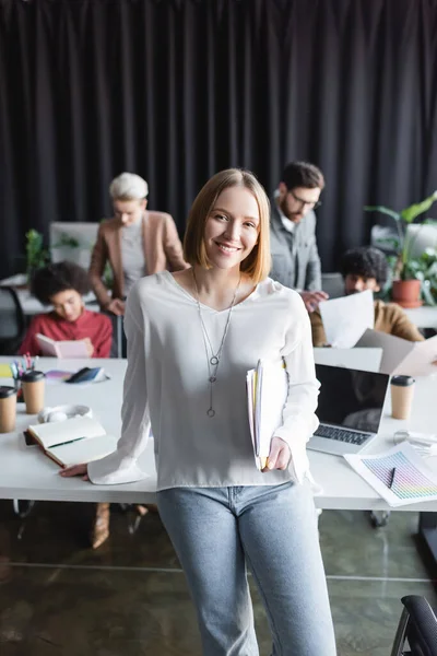 Glückliche Frau mit Ordnern, die in die Kamera in der Nähe multiethnischer Teams schauen, die an verschwommenem Hintergrund arbeiten — Stockfoto