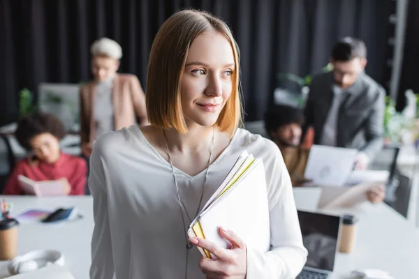 Feliz mujer sosteniendo carpetas cerca borrosa equipo multiétnico en agencia de publicidad - foto de stock