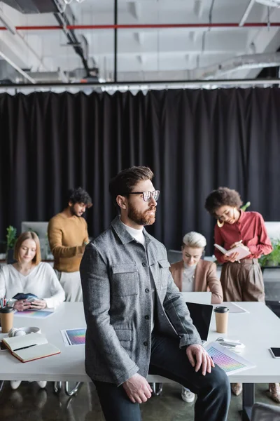 Businessman in eyeglasses looking away while multiethnic team working in advertising agency — Stock Photo