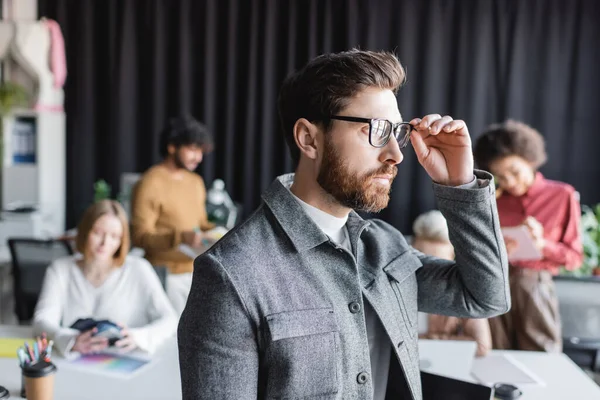 Bearded man adjusting eyeglasses and looking away near blurred interracial team in ad agency — Stock Photo