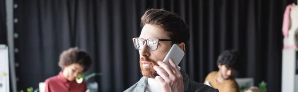 Менеджер в окулярах розмовляє на мобільному телефоні біля розмитих міжрасових колег в рекламному агентстві, банер — стокове фото