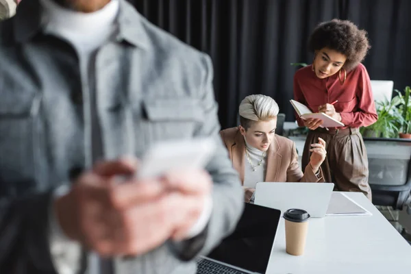 Uomo sfocato utilizzando il telefono cellulare vicino alle donne interrazziali che lavorano in agenzia pubblicitaria — Foto stock