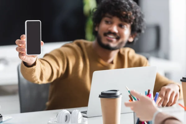 Fröhlicher indischer Mann hält Smartphone mit leerem Bildschirm neben Kollege auf verschwommenem Vordergrund — Stockfoto