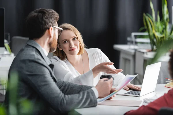 Smiling woman pointing with finger near laptop and colleague in ad agency — Stock Photo