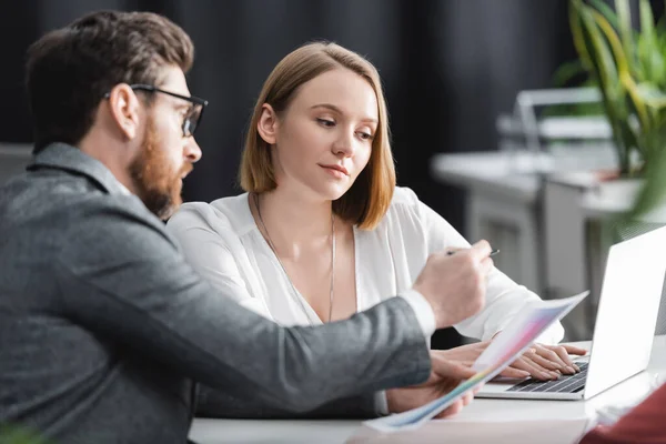 Blurred advertising manager showing paper to woman typing on laptop in office — Stock Photo