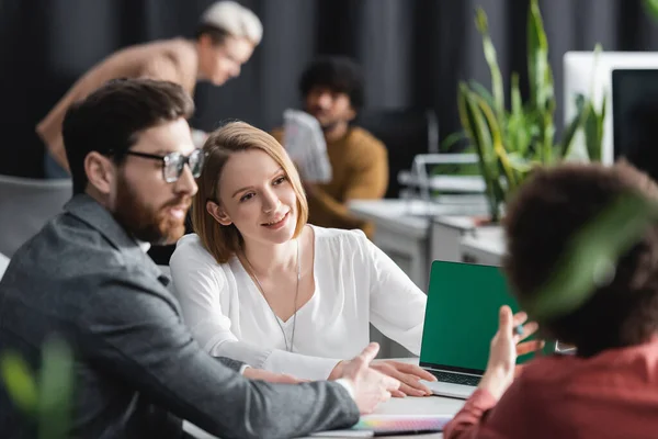 Woman smiling near multiethnic colleagues and laptop with green screen in ad agency — Stock Photo