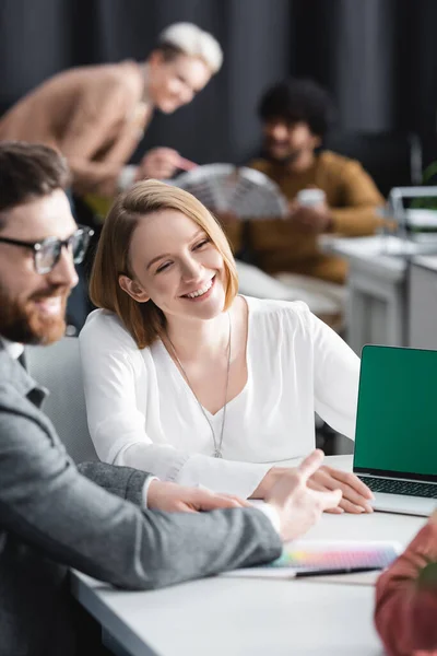 Счастливая женщина улыбается рядом с коллегами и ноутбук с зеленым экраном в рекламном агентстве — стоковое фото