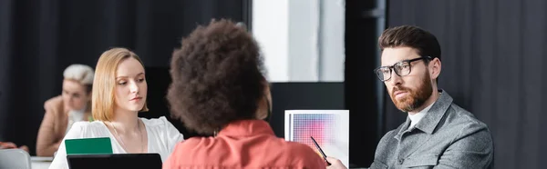 Homem em óculos apontando para amostras de cores perto de designers inter-raciais na agência de publicidade, banner — Fotografia de Stock