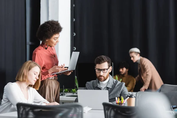 Donna afroamericana che punta al computer portatile vicino a colleghi multietnici che lavorano in agenzia pubblicitaria — Foto stock