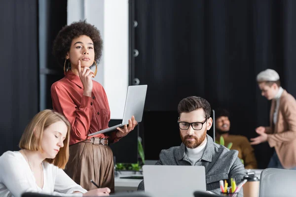 Mulher africana americana com laptop pensando perto de colegas que trabalham na agência de publicidade — Fotografia de Stock