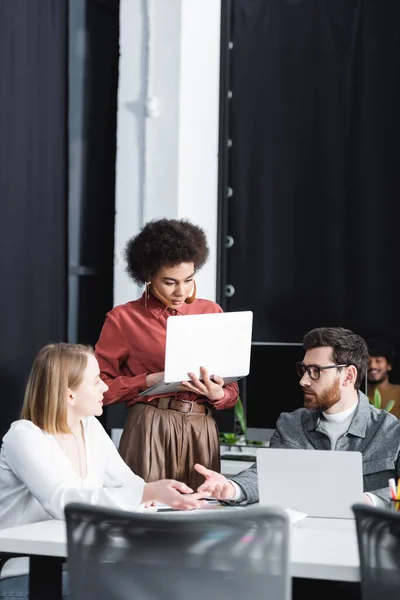 Mujer afroamericana usando laptop cerca de colegas hablando en agencia de publicidad - foto de stock