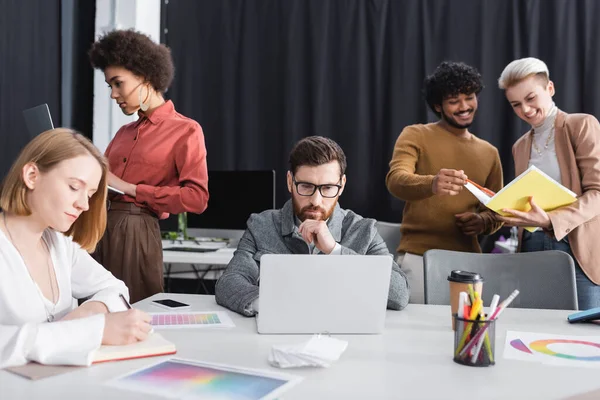 Homem em óculos pensando perto de laptop enquanto agentes de publicidade interracial trabalhando no escritório — Fotografia de Stock