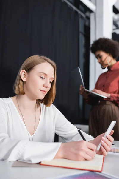 Donna d'affari che scrive nel taccuino vicino alla sfocata collega afroamericana in agenzia pubblicitaria — Foto stock