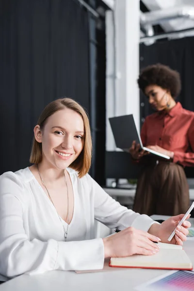 Manager pubblicitario con smartphone e notebook vicino al collega afroamericano su sfondo sfocato — Foto stock