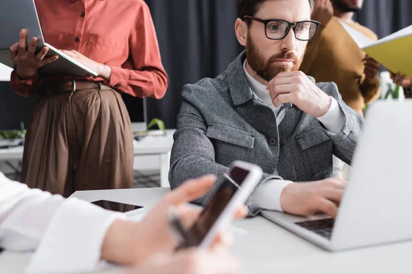 Pensive bearded man in eyeglasses working near laptop and blurred multicultural team in ad agency — Stock Photo