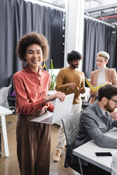 Joyeux agent de publicité afro-américain avec ordinateur portable regardant la caméra près de collègues multiculturels — Photo de stock