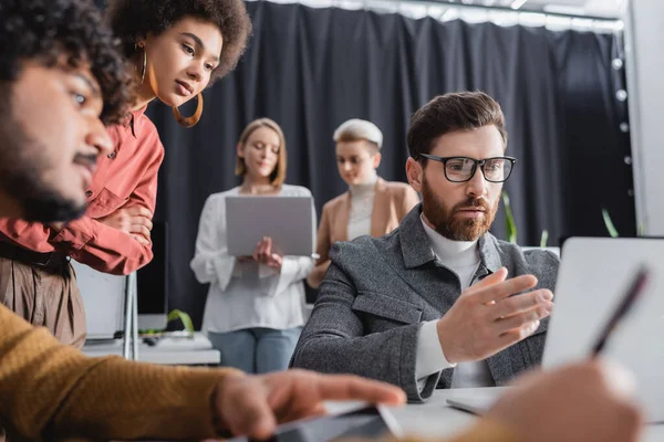 Pensativo hombre de negocios en gafas apuntando a la computadora portátil cerca de colegas multiculturales en la agencia de publicidad - foto de stock