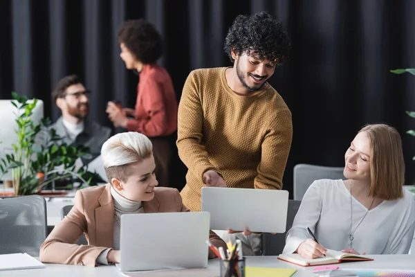 Hombre indio feliz mostrando el ordenador portátil a los colegas en la agencia de publicidad - foto de stock