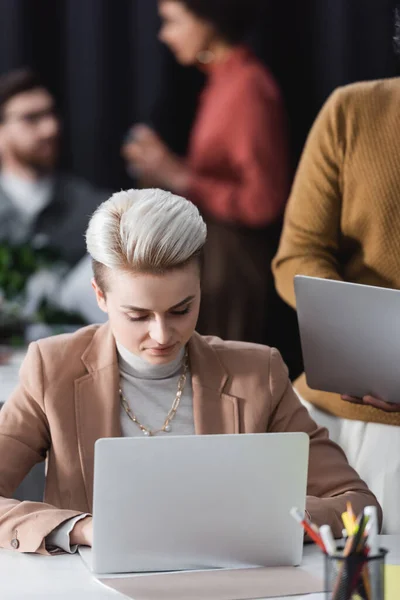 Femme blonde travaillant près d'un ordinateur portable et équipe floue dans l'agence de publicité — Photo de stock
