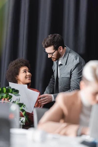 Feliz mulher afro-americana segurando papéis perto do homem em óculos e colega turvo na agência de publicidade — Fotografia de Stock