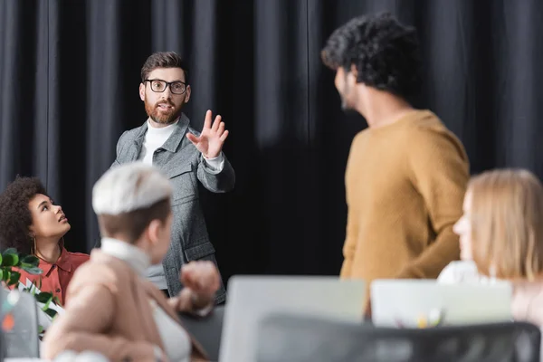 Advertising manager in eyeglasses pointing with hand near multicultural coworkers in office — Stock Photo