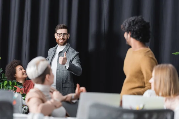 Cheerful man in eyeglasses showing thumb up near blurred multiethnic colleagues in ad agency — Stock Photo