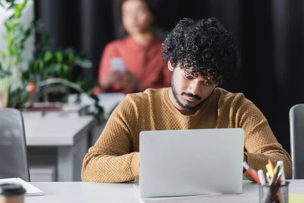 Curly indian advertising manager working on laptop near blurred african american woman — Stock Photo