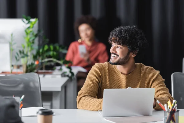 Happy indian advertising manager looking away near laptop and blurred african american colleague — Stock Photo