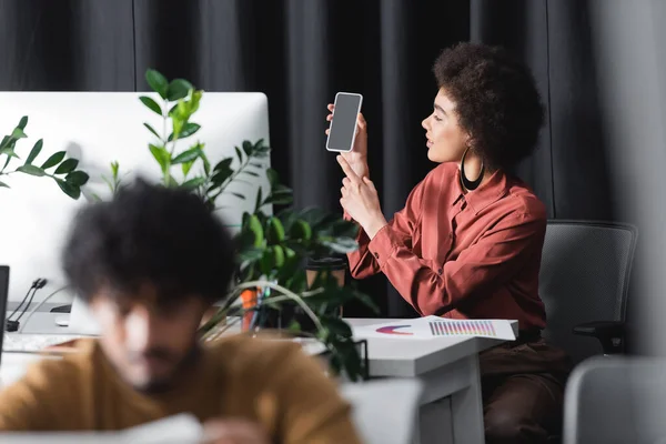 Mulher afro-americana apontando para smartphone perto de monitor de computador e colega indiano turvo — Fotografia de Stock