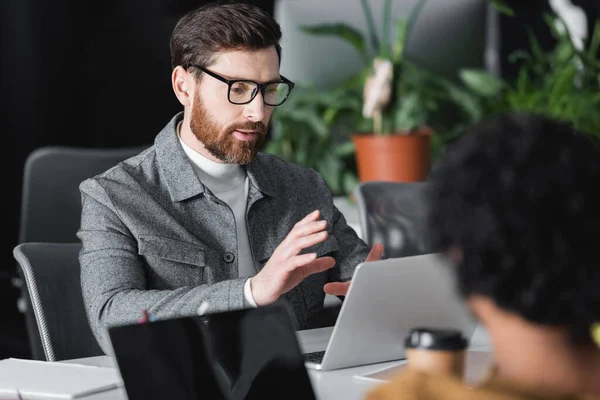 Hombre barbudo gesto cerca de la computadora portátil durante la videollamada en la agencia de publicidad - foto de stock